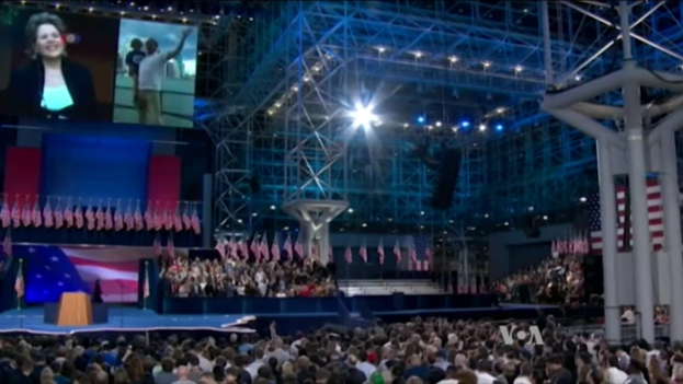 Große Menschenmenge vor einer Bühne mit einem Rednerpult, Mikrofon, Flaggen, Bildschirm und Lichtinstallationen, was an eine Präsidentschaftsdebatte erinnert.