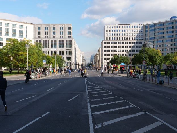 Eine belebte Stadtstraße in Berlin, Deutschland, mit Fußgängern und Radfahrern auf der Straße, hohen Gebäuden, Bäumen, Laternen und Schildern unter einem bewölkten Himmel.