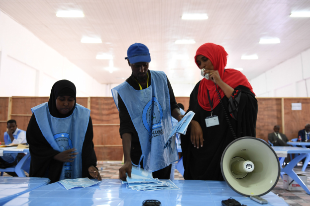 Eine Gruppe von Menschen steht um einen Tisch mit einem Megafon, Papieren und anderen Gegenständen an einer Wahllokal, während andere im Hintergrund auf Stühlen sitzen und stehen, unter beleuchteten Deckeninstallationen.