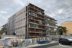 Ein großes Gebäude im Bau mit Gerüsten, umgeben von Bäumen, Straßenlaternen, Fahrzeugen, Mülltonnen und Werbetafeln unter einem bewölkten Himmel.