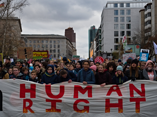Eine Gruppe von Menschen mit einer "Menschliche Rechte"-Fahne vor einer Menge mit Schildern und Fahnen, mit Bäumen, Gebäuden und Laternen im Hintergrund.