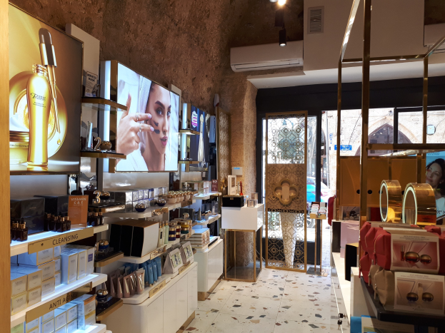 Innenansicht eines Kosmetikladens mit verschiedenen Produkten auf Gestellen, Wandbildschirmen, Deckenbeleuchtung, einer Klimaanlage und Glastüren, die eine Person, ein Auto und ein Gebäude draußen zeigen.