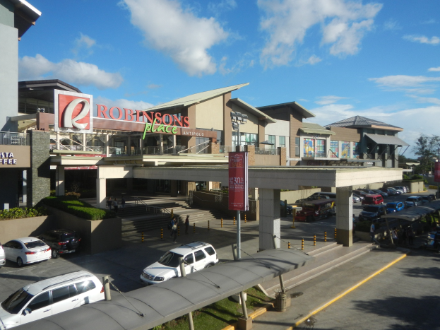 Ein belebtes Einkaufszentrum mit Gebäuden, Geschäften, Fahrzeugen, Fußgängern, Straßenmöblierung, Grünflächen und einem bewölkten Himmel.