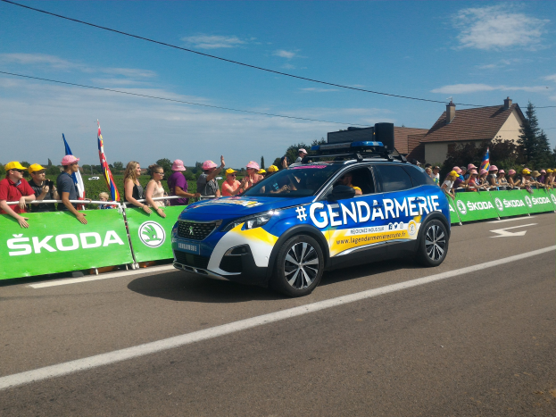 Polizeiauto fährt an einer Menge vorbei, die neben Bäumen und Gebäuden steht, mit einer Fahne auf dem Auto und Text, der unten 'Gendarmerie de la Tour de France' lautet.