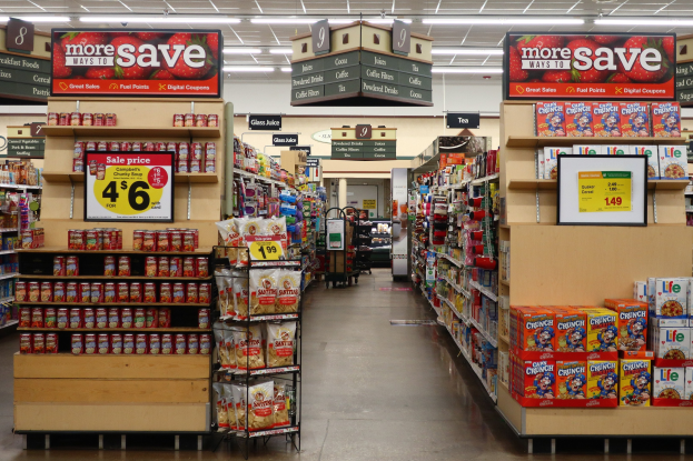 Ein Convenience-Store-Interieur mit Regalen und Bodenbestand mit verschiedenen Lebensmitteln, Deckenbeleuchtung und Informationsschildern im Hintergrund.