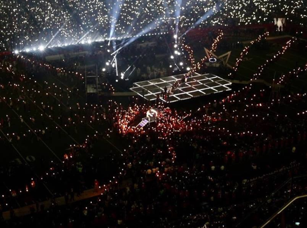 Eine große Menschenmenge in einem hell erleuchteten Stadion bei Nacht, mit einer Person, die auf einer Bühne in der Mitte während der Olympischen Schlussfeier performs.