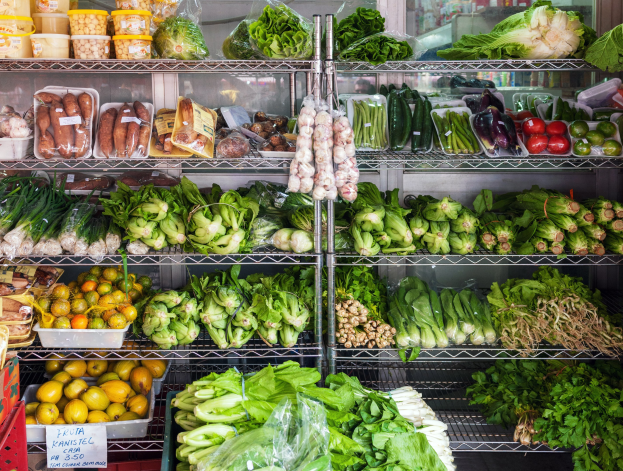 Frische Obst- und Gemüseauslage in einem Supermarkt mit Gemüse, Obst, verpackten Artikeln, Kartons, einer Schautafel und einem Glasfenster im Hintergrund.