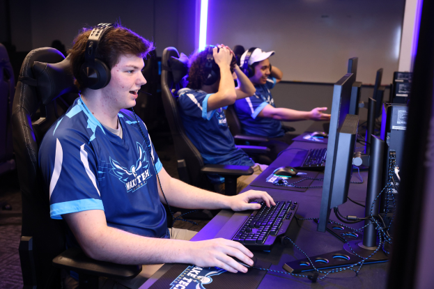 Gruppe junger Männer mit Headsets, die ein Videospiel auf Computern mit Monitoren, Tastaturen und Mäusen an einem Schreibtisch spielen, vor einer beleuchteten Wand.