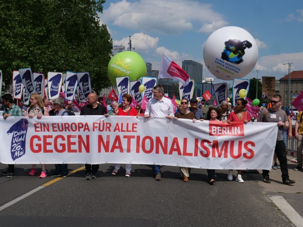 Eine Gruppe von Menschen marschiert bei einer Demonstration gegen Nationalismus in Berlin, trägt ein Banner, Fahnen und Luftballons, mit Bäumen, Gebäuden und einem bewölkten Himmel im Hintergrund.