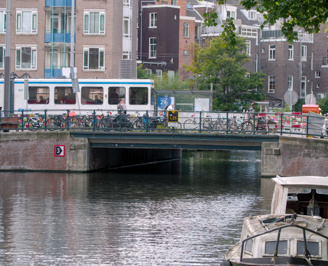 Ein Boot fährt auf einem Fluss neben einer Brücke mit Fahrrädern und Menschen darauf vorbei, mit Gebäuden, Bäumen und einem klaren blauen Himmel im Hintergrund.