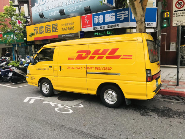 Gelbes DHL-Lieferfahrzeug steht am Straßenrand neben Häusern, Bäumen, Schildern, Strommasten, Ampeln und einem Fußgänger auf dem Gehweg.