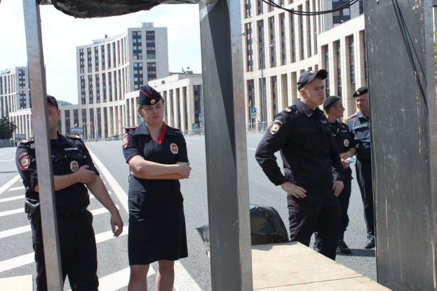 Eine Gruppe von Polizisten in Uniform auf einer Straße mit Pfählen und Drähten im Vordergrund, Gebäude, Bäume und einen klaren blauen Himmel im Hintergrund.