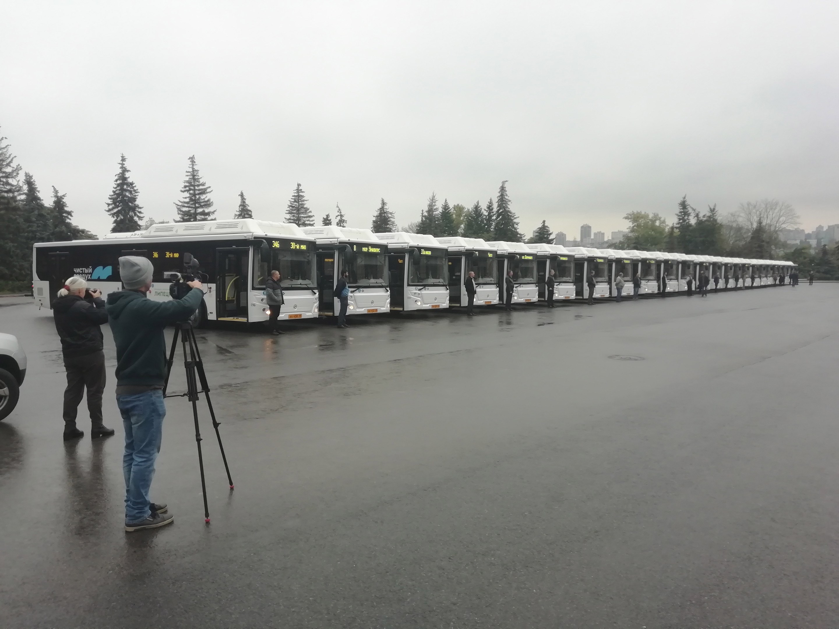 Eine Gruppe von Menschen, die eine Reihe von Bussen auf einer Straße mit Bäumen, Gebäuden und einem klaren blauen Himmel fotografieren