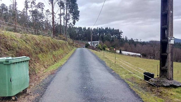 Grüner Mülltonnen am Rande einer Straße umgeben von Gras, Pflanzen, Bäumen, Polen, Drähten und einem Zaun, mit einem bewölkten Himmel im Hintergrund.