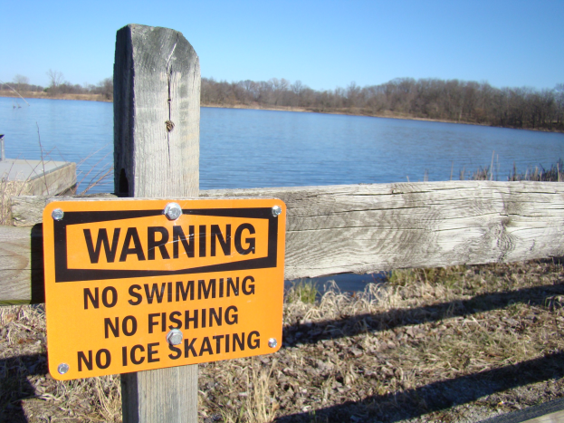 Warnschild an einem hölzernen Zaun in der Nähe eines Sees, mit Bäumen und einem klaren blauen Himmel im Hintergrund, auf dem steht "Kein Schwimmen, Kein Angeln, Kein Eislaufen".