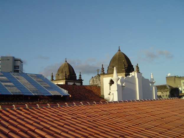 Stadtpanorama mit Gebäuden im Vordergrund und einem blauen Himmel im Hintergrund, das Solarpanels auf einem Dach zeigt, was den Einsatz erneuerbarer Energien anzeigt.
