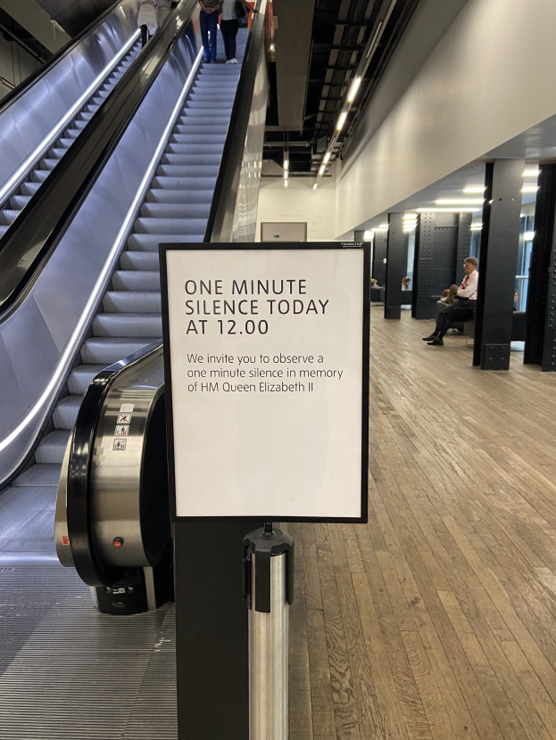 Eine Rolltreppe in einem Flughafen mit einem Schild, auf dem "Eine Minute Stille heute" steht, sowie einige Personen darauf und Lampen an der Decke im Hintergrund.