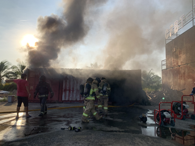 Feuerwehrleute in Helmen arbeiten daran, ein Gebäude Feuer zu löschen, mit dicken Rauch, während ein anderes Gebäude und Bäume in der Nähe sichtbar sind.