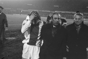 Eine Gruppe von Männern steht zusammen auf einem Fußballfeld, sieht besorgt aus und hat ein Stadion im Hintergrund, dargestellt in Schwarz-Weiß.