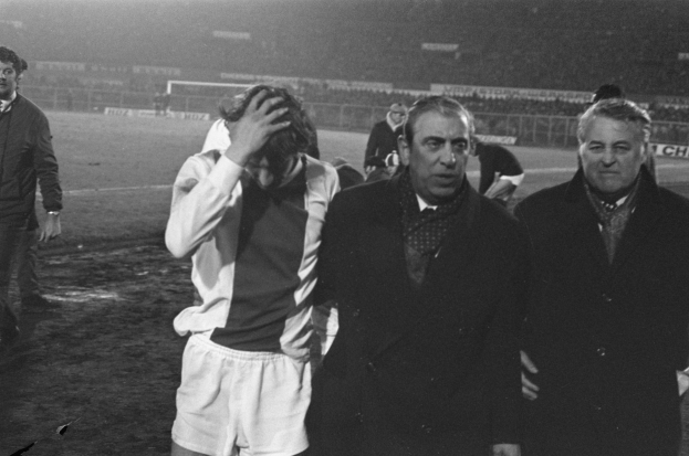 Eine Gruppe von Männern steht zusammen auf einem Fußballfeld, sieht besorgt aus und hat ein Stadion im Hintergrund, dargestellt in Schwarz-Weiß.