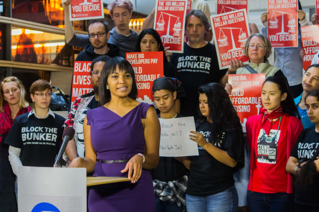 Eine Black Lives Matter-Aktivistin spricht an einem Rednerpult mit Mikrofon zu einer Menge mit Schildern, mit Lichtern und Gegenständen im Hintergrund.