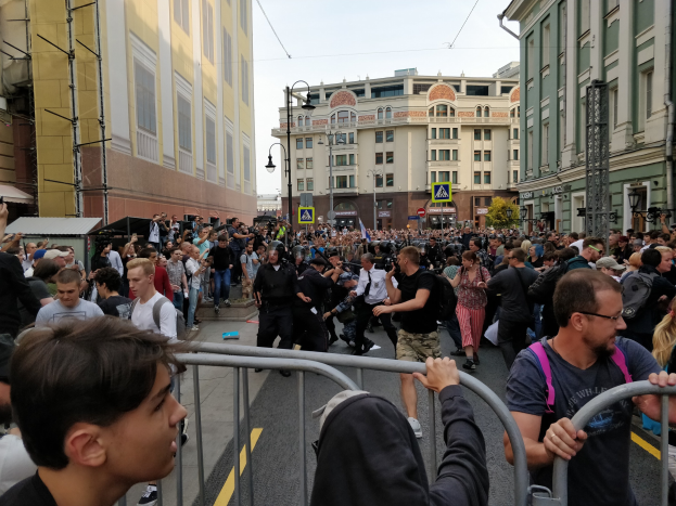 Eine große Menschenmenge steht hinter Metallbarrieren auf einer Straße, umgeben von Gebäuden, Bäumen und Laternen, wobei einige Kameras halten.