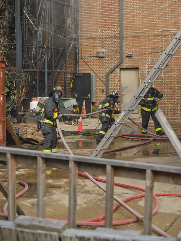 Feuerwehrleute in Helmen arbeiten daran, ein Gebäude Feuer zu löschen, umgeben von Equipment und einem Metallzaun, mit einem Baum und Himmel im Hintergrund.