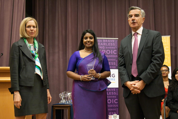 Zwei Frauen und ein Mann auf einer Bühne bei den Women's Leadership Awards 2016; die Frau in der Mitte hält einen Award in einem lila Kleid, der Mann rechts trägt einen schwarzen Blazer, ein weißes Hemd und eine rosa Krawatte, mit einem Podium und Mikrofon links und Awards auf einem Tisch davor.
