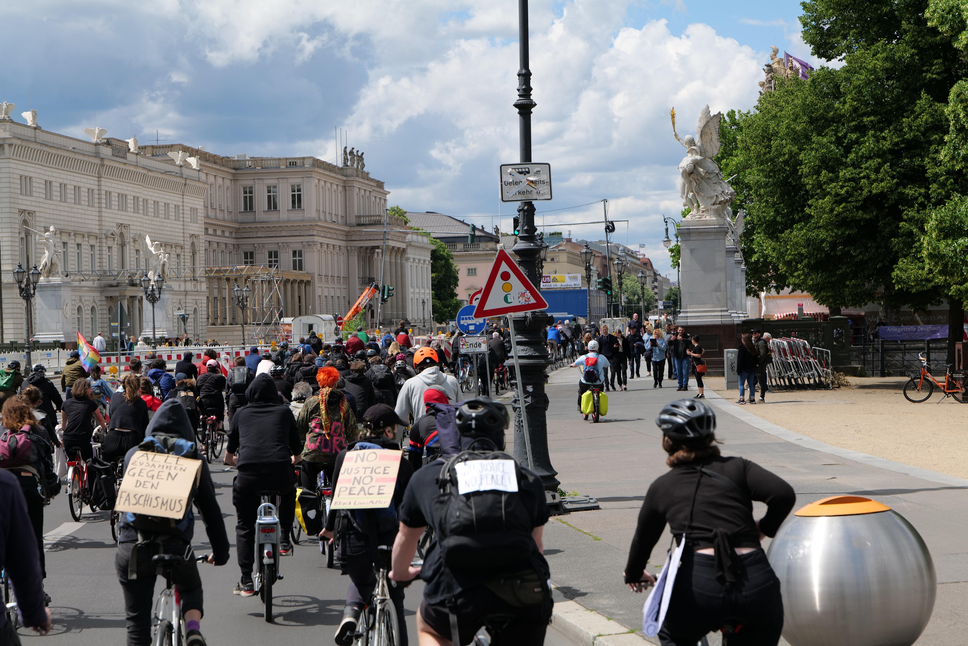 Eine große Gruppe von Radfahrern fährt eine von hohen Gebäuden gesäumte Stadtstraße entlang, einige tragen Helme und haben Taschen dabei, andere halten Schilder; Pfosten mit Schildern und eine Statue sind im Hintergrund zu sehen, zusammen mit Bäumen und einem bewölkten Himmel.