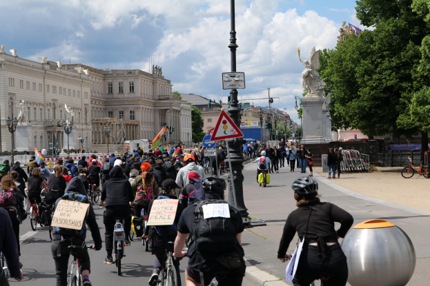 Eine große Gruppe von Radfahrern fährt eine von hohen Gebäuden gesäumte Stadtstraße entlang, einige tragen Helme und haben Taschen dabei, andere halten Schilder; Pfosten mit Schildern und eine Statue sind im Hintergrund zu sehen, zusammen mit Bäumen und einem bewölkten Himmel.
