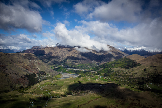 Eine Totalenaufnahme einer bergigen Landschaft in Queenstown, Neuseeland, mit saftig grünem Gras, verstreuten Bäumen, einer gewundenen Straße und einem Himmel voller weißer, flauschiger Wolken.