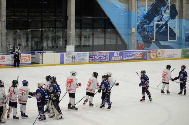 Gruppe junger Menschen in Helmen und Hockey-Ausrüstung, die auf einer Indoor-Eisbahn mit Bannern, Säulen und Glaswänden im Hintergrund Eishockey spielen.