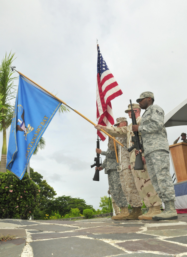 Eine Gruppe von Männern in Militäruniformen mit Waffen in der Hand, die neben einem Podium mit einem Mikrofon vor einem Hintergrund aus Bäumen, Pflanzen und einem klaren blauen Himmel stehen.