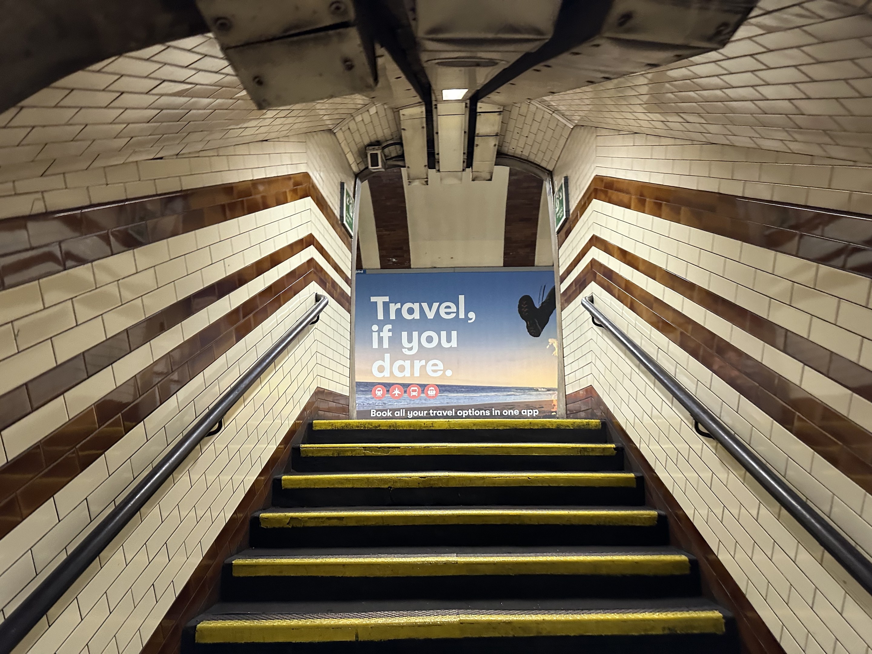 Eine Treppe in einer U-Bahn-Station mit einem 'Reise, wenn du wagst'-Schild oben, flankiert von Geländern und sichtbaren Stationswänden.