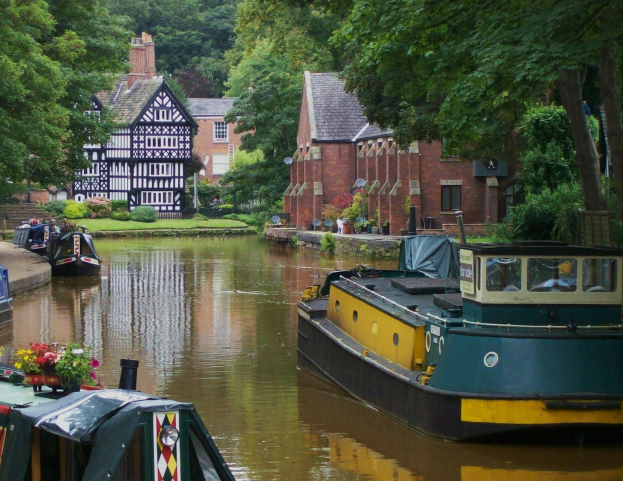Kanal mit mehreren Booten, die an seinem Rand vertäut sind, umgeben von grünem Gras, Pflanzen und Bäumen, mit Gebäuden mit Fenstern im Hintergrund.