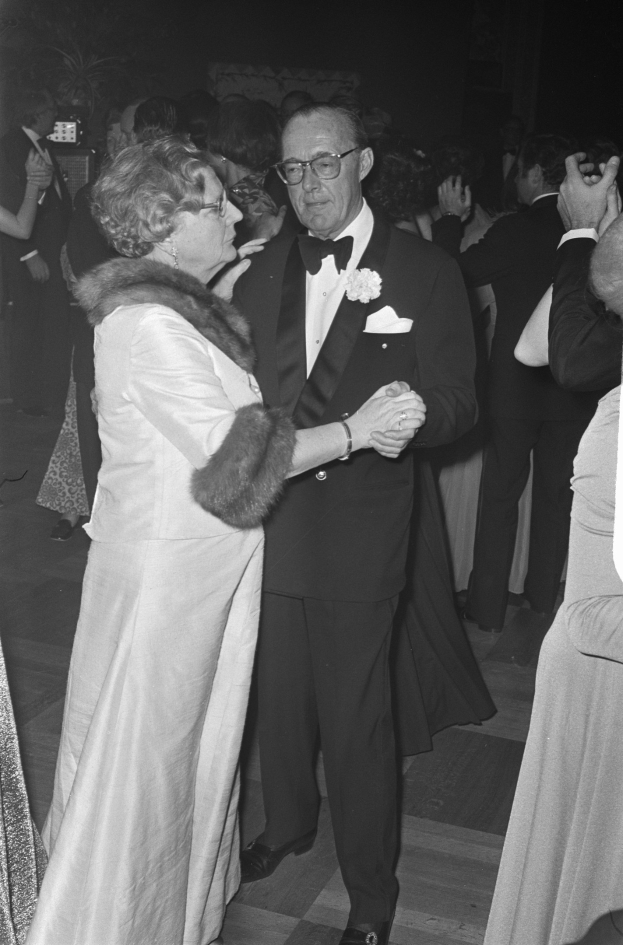 Ein Mann in einem schwarzen Blazer, weißem Hemd und schwarzen Hosen tanzt mit einer Frau in einem eleganten Kleid auf einer Party, umgeben von anderen Menschen und Kameras im dunklen Hintergrund.