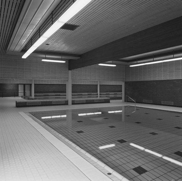 Indoor-Schwimmbad mit klarem Wasser, gefliestem Boden und Wänden, Deckenbeleuchtung und einigen umliegenden Gegenständen in einem großen, ruhigen Gebäude.