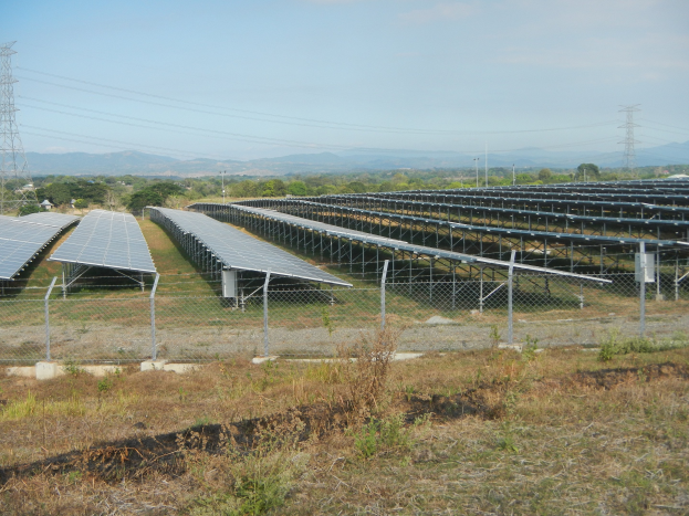 Luftaufnahme eines Solarpanelfelds, das von einem Zaun umgeben ist, mit Gras und Pflanzen im Vordergrund, Bäumen und Übertragungsmasten im Hintergrund und Hügeln unter einem klaren blauen Himmel.