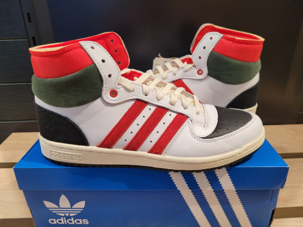Weiße, rote, grüne und schwarze Adidas Originals Top Ten Hi Schuhe auf einer blauen und braunen Kartonschachtel auf einem cremefarbenen Tisch vor einer schwarzen Wand.