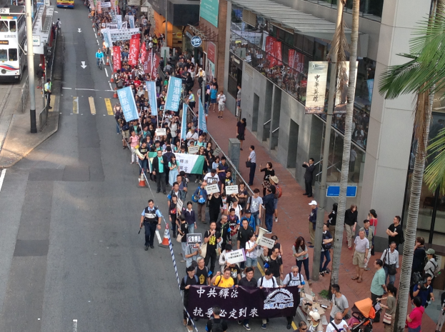 Eine große Gruppe von Menschen marschiert auf einer Straße in Hong Kong, wobei Protestschilder gehalten werden, und es sind Bäume, Glasfassaden, Fahrzeuge und Schilder zu sehen.
