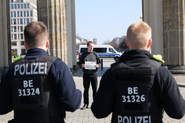 Drei Polizeibeamte stehen vor einem Gebäude, wobei ein Mann in der Mitte ein Papier hält und von Säulen und Fahrzeugen auf der Straße dahinter flankiert wird.