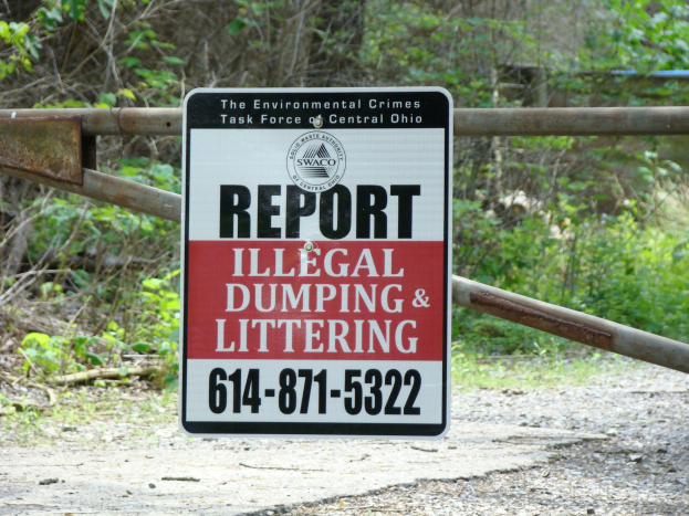 Ein Schild am Straßenrand trägt die Aufschrift "Melde illegalen Müll und Littering" mit Bäumen und Pflanzen im Hintergrund und einer Tafel mit Text im Vordergrund.