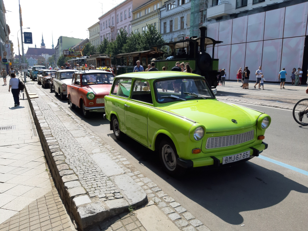 Ein grünes Auto fährt auf einer Straße mit hohen Gebäuden, Fußgängern auf einem Gehweg, einem Radfahrer, Bäumen und einem klaren blauen Himmel im Hintergrund.