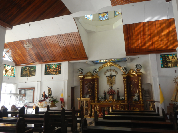 Inneneinrichtung einer Kirche mit Holzlehnen, Statuen, Blumenbouquets, Flaggen, Fotorahmen an den Wänden und Buntglasfenstern, mit Deckenleuchten, die eine gemütliche Atmosphäre schaffen.