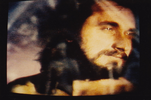 Ein Mann mit Bart, Schnurrbart und langen dunklen Haaren, der eine weiße Hemd trägt, schaut direkt in die Kamera auf einem TV-Bildschirm mit dunklem Hintergrund.