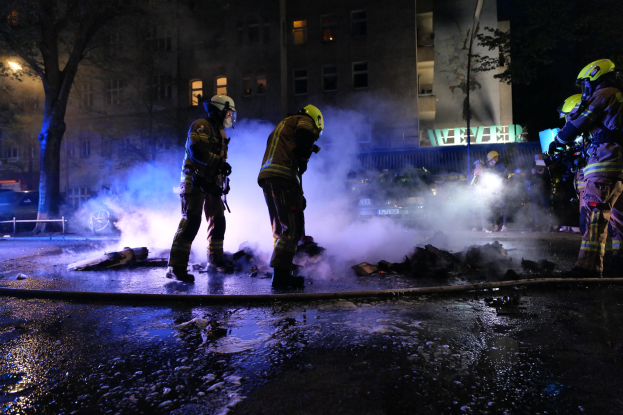Feuerwehrleute in Helmen stehen nachts auf einer Straße mit Rauch, der aus einer Leitung auf der Straße aufsteigt, und halten Gegenstände in den Händen, während Gebäude, Bäume und Fahrzeuge im Hintergrund zu sehen sind, mit einer Tafel auf der rechten Seite.