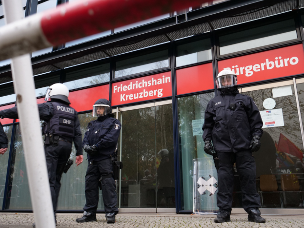 Polizeibeamte in Helmen und Handschuhen stehen vor einem Gebäude mit Glaswänden und -türen, neben einem Pfahl, mit Schildern, die "Friedrichshain Kreuzberg" im Hintergrund zeigen.
