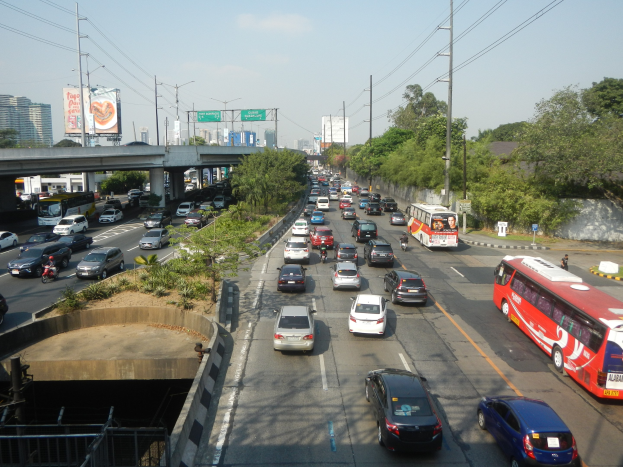 Vollgestopfte Stadtstraße mit Autos, Bussen, Strommasten, Bäumen, Gebäuden, Werbetafeln und einer Brücke unter einem bewölkten Himmel.