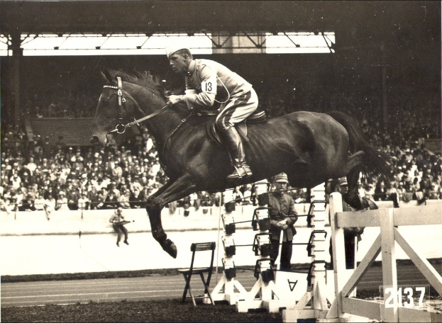 Ein Mann in weißer Kleidung reitet auf einem braunen Pferd über ein Hindernis in einer Arena, während Zuschauer zusehen.