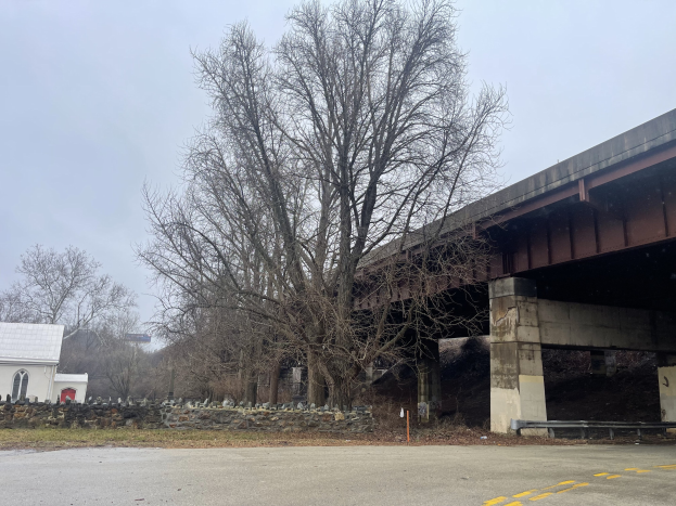 Brücke in Reparatur mit verstreuten Werkzeugen und Materialien, Baum in der Mitte umgeben von Steinen, Gras, einem Haus mit Dach, einem Schild, einem Straßenpfahl, einer Gruppe von Bäumen und einem bewölkten Himmel.